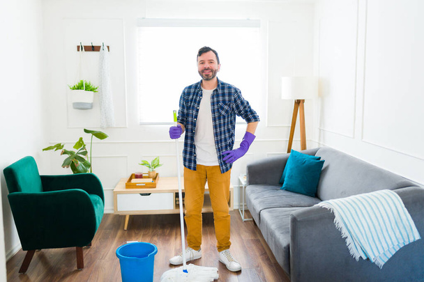 Retrato de um homem caucasiano feliz sorrindo olhando para a câmera enquanto se prepara para limpar sua sala de estar usando uma esfregona - Foto, Imagem