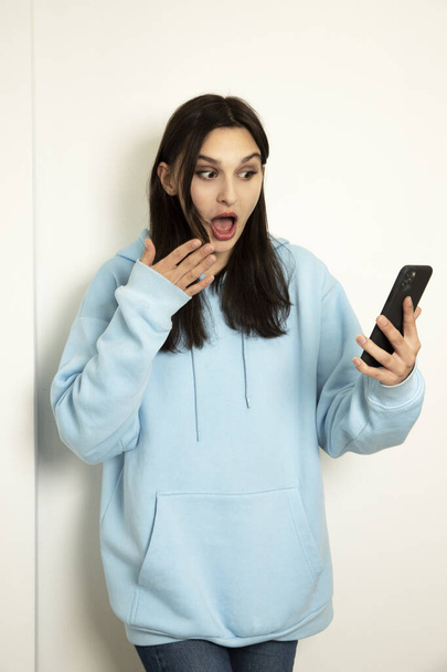 emotional girl with a phone in a blue jacket on a light background. The model is shocked. - Foto, Imagem