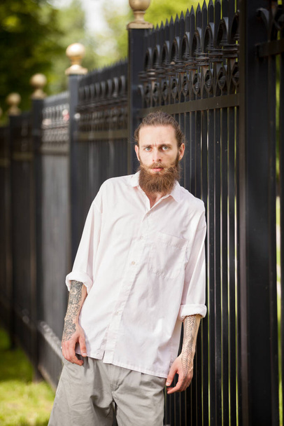 Hipster mit langem Bart posieren außerhalb der Stadt neben einem Zaun. Stil und Vielfalt - Foto, Bild