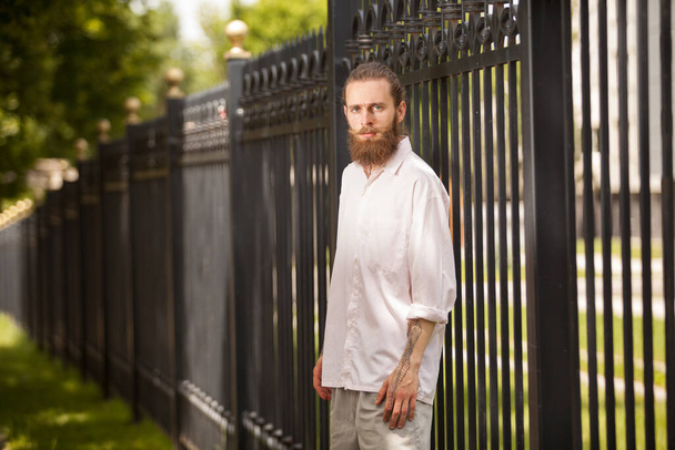 Ein bärtiger Hipster posiert neben einem Zaun. Stil und Vielfalt - Foto, Bild