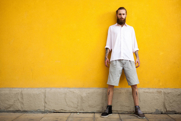 Cool hipster barbudo tatuado en la pared amarilla posando al aire libre
 - Foto, Imagen
