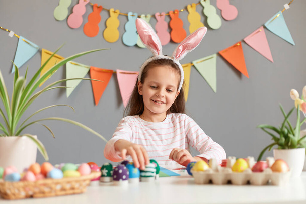 Portrait of satisfied delighted little girl wearing rabbit ears sitting at table painting eggs preparing for Easter, waiting for holiday, against gray wall with decorations - Fotó, kép