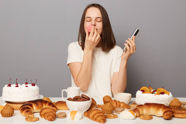 Photo of sugar addicted woman wearing white T-shirt sitting at table smells macaroons, impossible to resist eating dessert, has good appetite, enjoys homemade bakery isolated over gray background - Foto, Imagem