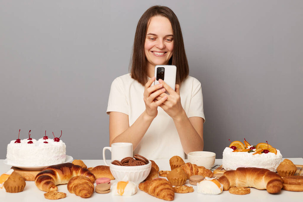 Delighted smiling cheerful millennial woman with brown hair wearing white T-shirt sitting at table chatting online via cell phone, enjoys eating desserts, isolated over gray background - Фото, изображение
