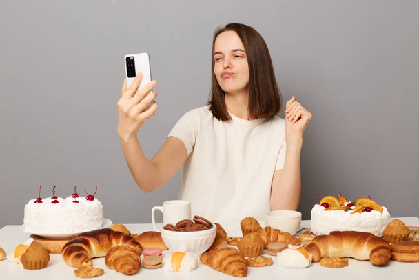 Beautiful positive woman with brown hair wearing white T-shirt sitting at table poses for selfie uses smartphone shows yummy desserts, pout lips, isolated over gray background - Fotó, kép