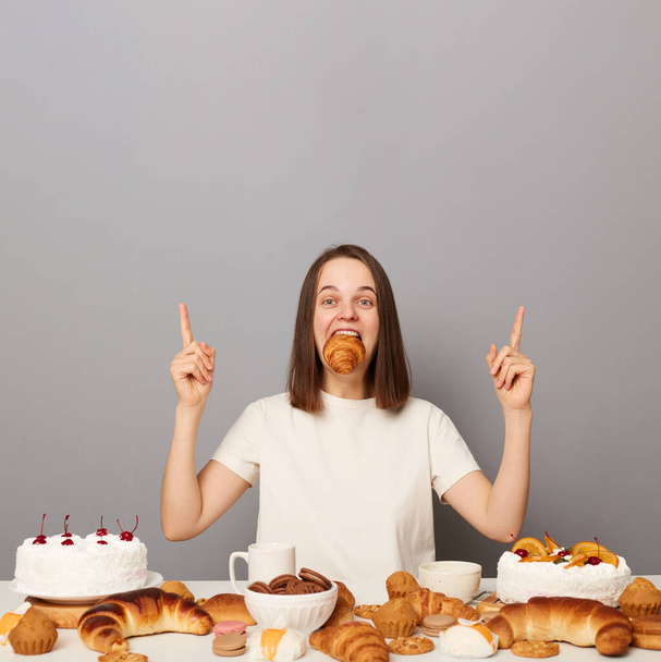 Portrait of excited woman with brown hair in white T-shirt sitting at table isolated over gray background, posing among big variety of desserts with full mouth of cookies, pointing up at copy space. - Zdjęcie, obraz