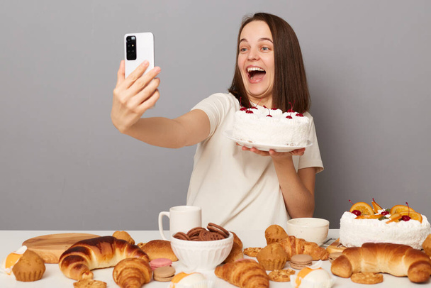 Portrait of amazed happy beautiful woman with brown hair wearing white T-shirt sitting at table isolated over gray background, celebrating birthday, talking selfie with cake. - Foto, afbeelding