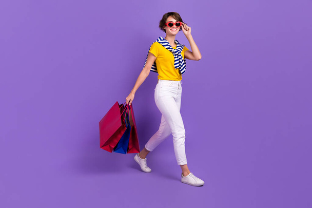 Full length photo of positive pretty lady wear bright clothes go empty space carry packages packs isolated on purple color background. - Photo, image