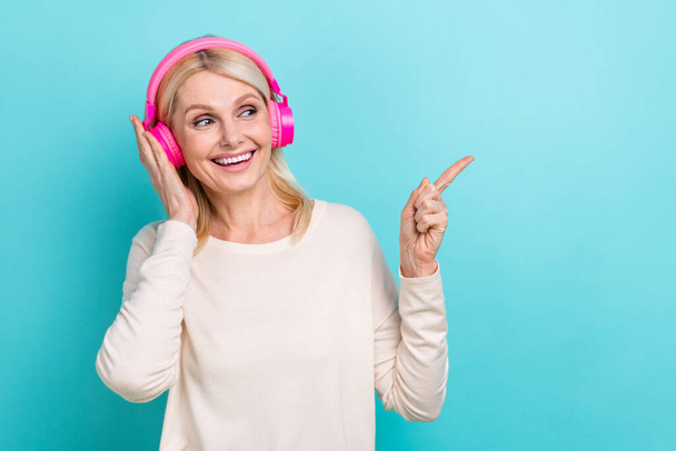 Photo of funky grandmother wear white shirt direct finger empty space touch sony earphones enjoy high quality sound isolated on blue color background. - 写真・画像