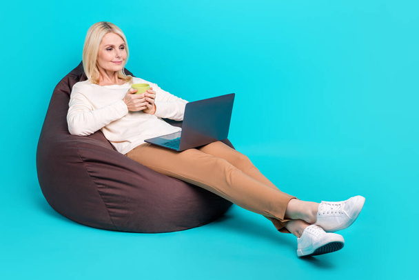 Full length photo of businesswoman senior sit pouf hold cup fresh coffee morning breakfast watch series netbook isolated on blue color background. - Foto, afbeelding