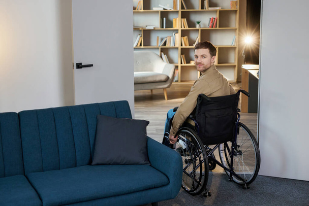 Een knappe jongeman in een rolstoel beweegt zich door het appartement, went aan een nieuw leven. Herstel na letsel of ziek eenzaam thuis. - Foto, afbeelding