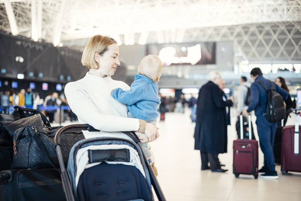 Moederdier reizen met zijn baby baby jongen kind, wandelen, duwen kinderwagen en bagage kar op luchthaven terminal station. Reizen met kind concept - Foto, afbeelding