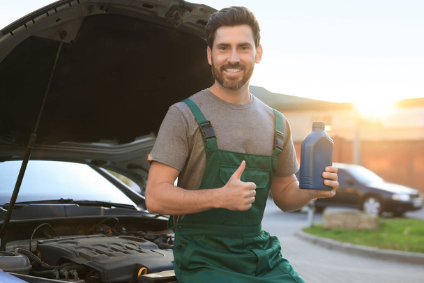 Smiling worker holding blue container of motor oil and showing thumbs up near car outdoors - Photo, Image