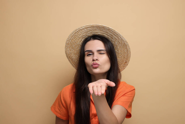 Beautiful young woman in straw hat blowing kiss on beige background - Fotoğraf, Görsel
