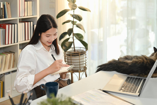 Mosolygó fiatal nő kisvállalkozás vállalkozó ül otthon irodai íróasztal és írási nyilvántartást a napi tervező. - Fotó, kép