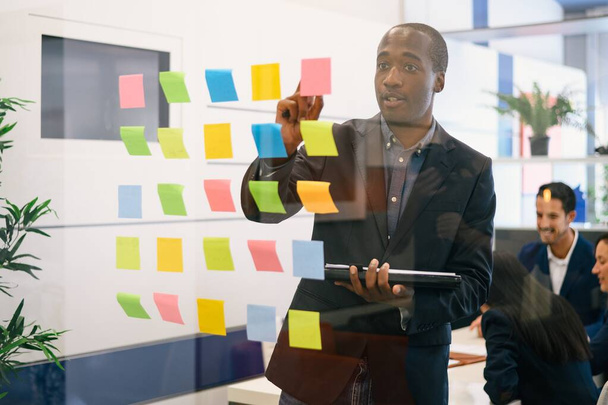 Zaměřený afroamerický podnikatel ve formálním obleku stojící u skleněné stěny s barevnými samolepícími poznámkami a čtením informací během setkání s kolegy v konferenční místnosti - Fotografie, Obrázek
