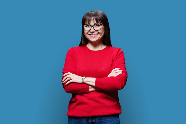 Confident middle aged woman with crossed arms in red casual jumper looking at camera on blue studio background. - Фото, изображение