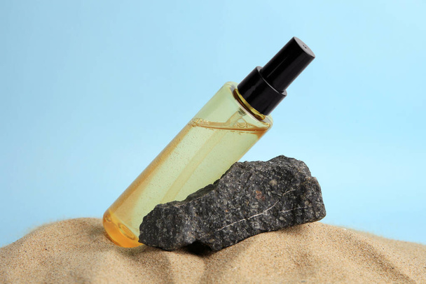 Botella con suero y piedra sobre arena sobre fondo azul claro, primer plano. Producto cosmético - Foto, Imagen
