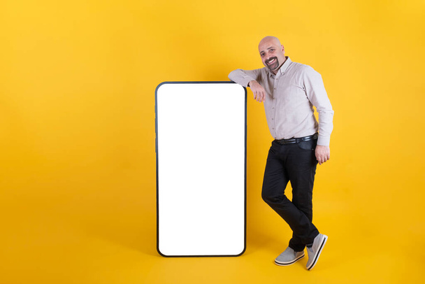 Διαφήμιση εφαρμογών για κινητά, άνθρωπος που γέρνει μεγάλο smartphone για διαφήμιση εφαρμογών για κινητά. Πλήρες μήκος του σώματος του χαρούμενος χαμογελαστός μεσήλικας άνδρας κοντά στο μεγάλο τεράστιο λευκό κενό κινητό τηλέφωνο οθόνη mock up. Αντιγραφή χώρου. - Φωτογραφία, εικόνα
