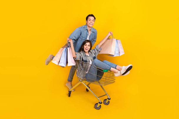 Full length photo of adorable excited best friends dressed denim rising bargains rising cart isolated yellow color background. - Fotoğraf, Görsel
