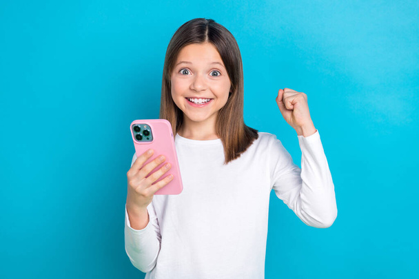 Photo portrait of cute little girl hold telephone raise fist like comment dressed stylish white clothes isolated on blue color background. - Φωτογραφία, εικόνα