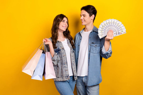 Portrait of two satisfied glad people hold boutique bags dollar banknotes look each other isolated on yellow color background. - Фото, изображение