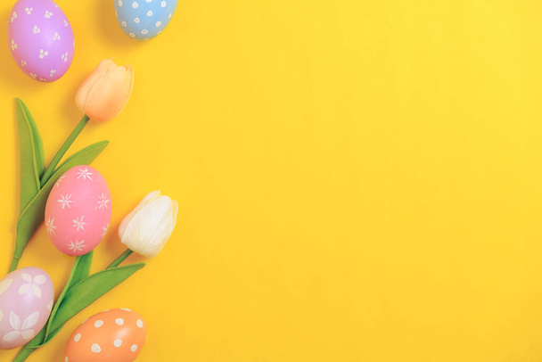Happy Easter holiday greeting card concept. Colorful Easter Eggs and spring flowers on yellow background. Top view, flat lay, copy space. - Foto, imagen