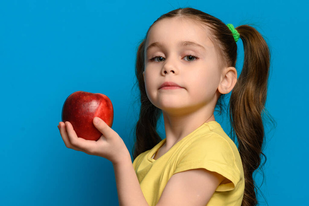 Το κορίτσι στέκεται στο προφίλ σε μπλε φόντο και κρατά ένα μεγάλο κόκκινο μήλο, αντιγραφή χώρο και παιδικές τροφές στο ένα χέρι. - Φωτογραφία, εικόνα