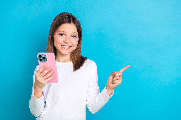 Photo of positive schoolgirl straight hairdo dressed white shirt hold smartphone directing empty space isolated on blue color background. - Foto, Bild