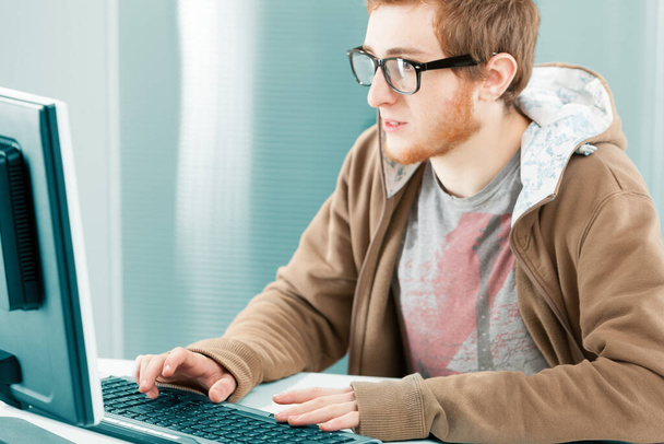 Bilgisayar ekranının önünde genç bir milenyum çalışanı. Siyah reçeteli gözlüklü, kahverengi kapüşonlu. Yazıyor, çok ilgili ve odaklanmış.. - Fotoğraf, Görsel