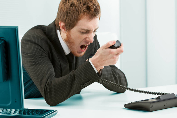 Controlling aggression in the work environment may be necessary. Man in a suit in the office yells violently at the telephone by grabbing the handset firmly and pouring all his anger into it by shouti - 写真・画像