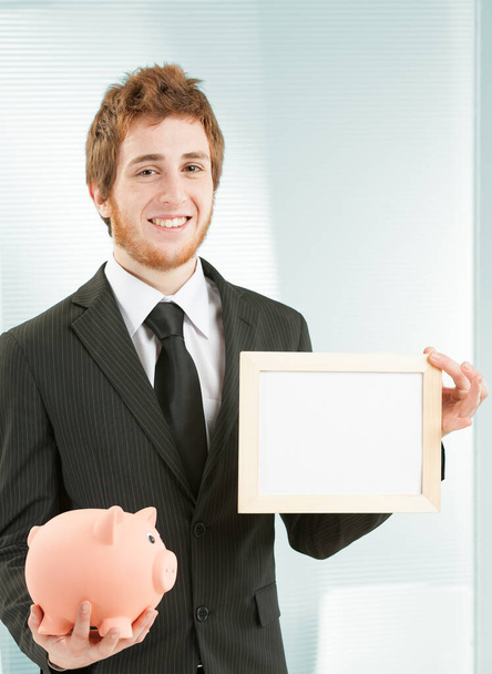 Copyspace από την έννοια της εξοικονόμησης. Νεαρός χαμογελαστός επιχειρηματίας κρατά μια λευκή πινακίδα διαθέσιμη για το γράψιμό σας στο ένα χέρι και ένα κουμπαρά που συμβολίζει τα χρήματα και τις αποταμιεύσεις στο άλλο. - Φωτογραφία, εικόνα