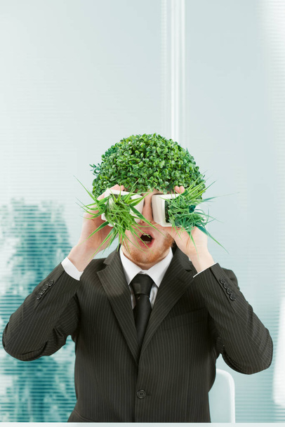 A clerk in an office makes a stunned face in a surreal mood. He has a bush for hair and looks through two pots of grass side by side as if through binoculars. 80s style in the mimicry. Black pinstripe - 写真・画像