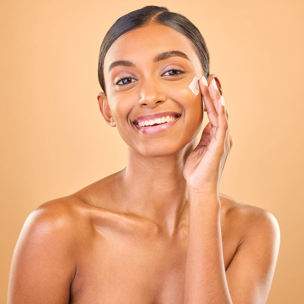 Face portrait, skincare and woman with cream in studio isolated on a brown background. Dermatology smile, cosmetics and happy Indian female model apply lotion, creme and moisturizer for healthy skin - Foto, imagen
