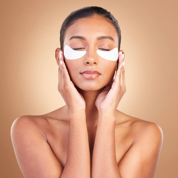 Skincare, eye patch and touch with Indian woman in studio for facial, spa treatment and glow. Self care, cosmetics and hydration with female model on brown background for mask, product and youth. - Photo, Image