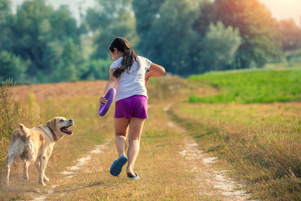A girl with a dog runs along a dirt path in the field - Fotoğraf, Görsel