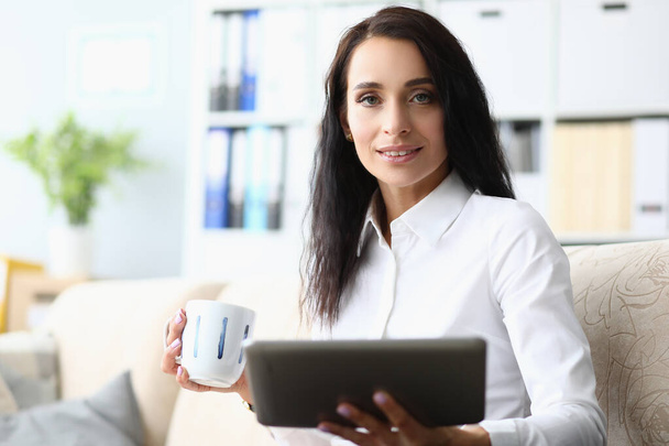 Πορτρέτο μιας όμορφης γυναίκας με tablet και κύπελλο στον καναπέ. Αιτήσεις για επιχειρήσεις και προγραμματισμός της εργάσιμης ημέρας - Φωτογραφία, εικόνα