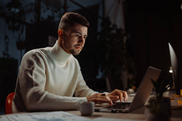 Homem focado vestindo roupas casuais elegantes usando laptop para o trabalho e tentando se apressar até o prazo enquanto sentado no espaço de escritório moderno. Funcionário do escritório respondendo e-mails, digitando à noite - Foto, Imagem