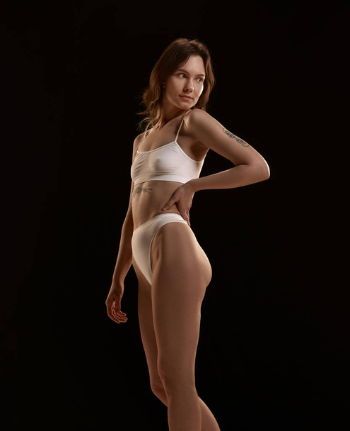 Attractive, young girl with slim, fit, healthy body posing in underwear against black studio background. Well-being and self-care. Concept of body and skin care, figure, fitness, health, wellness. - Fotoğraf, Görsel