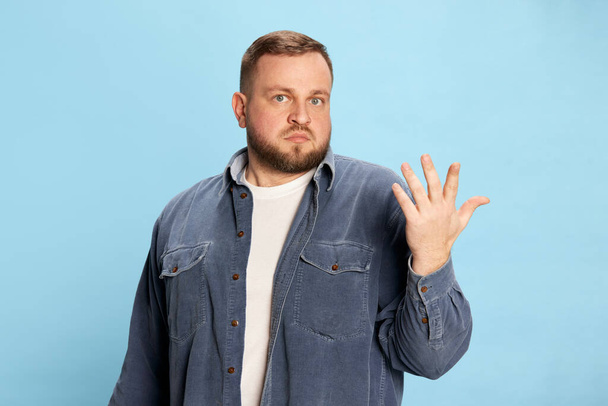 No idea, no clue. Good-looking man wearing jeans jacket spreading hand and looking at camera with face without understanding over blue background. Concept of emotions, facial expression, mood - Foto, imagen