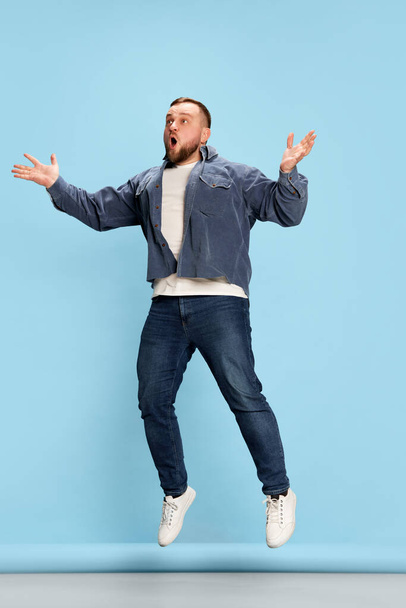 Bounce, levitation, happiness. Portrait of vivid emotional man wearing jeans and jumping with joyful face over blue background. Concept of bright emotions, facial expression, mood - Photo, image