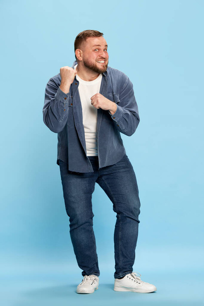Feeling of victory, win. Excited bearded man in denim costume showing clenches fists, smiling and looking away over blue background. Concept of bright emotions, facial expression, mood, success, ad - Foto, Imagen
