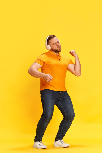 Dance, sing song, favourite melody. Exciting man wearing headphones listening music and dancing with hands over yellow background. Concept of music, hobby, human emotions, mood, ad - Фото, изображение