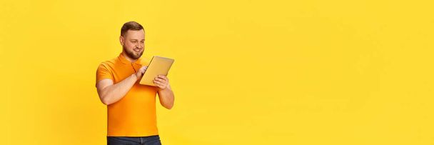 Verkoper, werk op afstand, online. Banner met een knappe man met behulp van digitale tablet en glimlachen met genieten gezicht geïsoleerd over gele achtergrond. Concept van het bedrijfsleven, verkoop, emoties, stemming, reclame - Foto, afbeelding