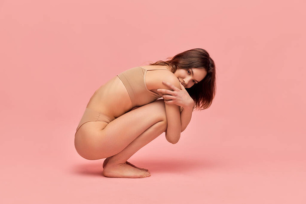 Tender femininity. Portrait of attractive, cute, young girl in beige underwear posing against pink studio background. Slim body. Concept of body and skin care, figure, fitness, health, wellness. - 写真・画像