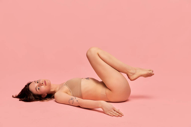 Relaxed and calm. Self-love and acceptance. Portrait of young girl in beige underwear posing against pink studio background. Concept of body and skin care, figure, fitness, health, wellness. - 写真・画像