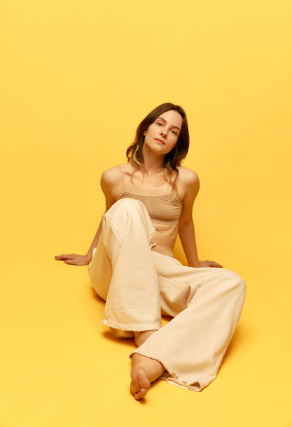 Beautiful young girl with relaxed calm face posing in top and pants against yellow studio background. Femininity. Concept of body and skin care, figure, fitness, health, wellness, beauty and fashion - Zdjęcie, obraz