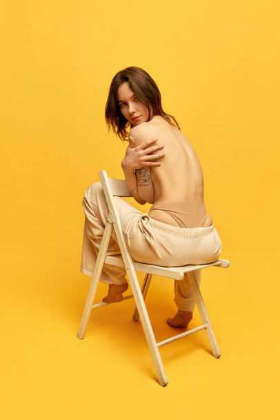 Rear view image of young slim girl with relief naked back sitting on chair in jeans, posing against yellow studio background. Concept of beauty, body and skincare, figure, fitness, health, wellness. - 写真・画像