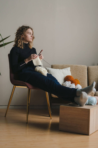 Jonge vrouw zittend in een gezellige fauteuil breiend met naalden en wollen draad zittend in een gezellige woonkamer. Handwerk, creatieve hobby of recreatie. - Foto, afbeelding