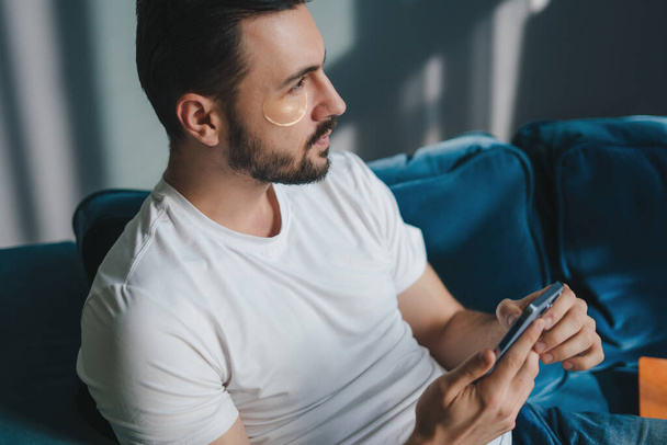 Young man wearing casual white t-shirt using mobile cell phone chat online sitting on sofa indoors at home on weekends. People leisure concept. Internet network - Zdjęcie, obraz